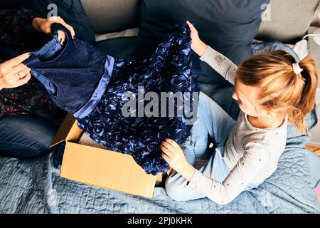 Mère et sa fille ouvrant la boîte avec robe commandée à la maison sur le canapé. Maman client en ligne tenant la robe. Service de livraison mode concept Banque D'Images