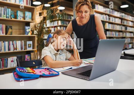 Professeur aidant à résoudre et à terminer la tâche. Ennuient une petite fille étudiante qui faisait ses devoirs à l'aide d'un ordinateur portable dans un club après l'école primaire. Retour à sc Banque D'Images