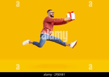 Jeune homme souriant dans des verres sautant haut tenant cadeau souriant isolé sur fond jaune. Banque D'Images