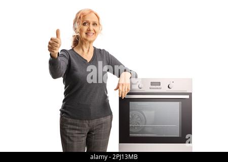 Femme mûre souriante se penchée sur un four et montrant les pouces isolés sur fond blanc Banque D'Images