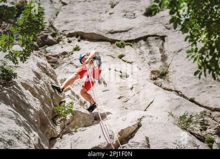 Grimpeur actif d'âge moyen dans un casque de protection tout en descendant du mur de roche de falaise à l'aide de cordes avec un dispositif de belay et un harnais d'escalade. Actif e Banque D'Images