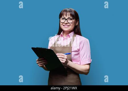 Femme d'âge moyen en tablier avec le presse-papiers posé sur fond bleu Banque D'Images