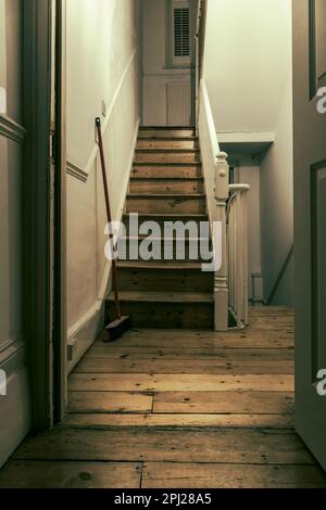 Une brosse penche contre un mur à l'extérieur d'une porte ouverte menant à dix marches d'un escalier en bois victorien. Banque D'Images