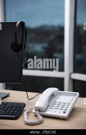 Téléphone fixe près d'un ordinateur moderne avec casque sur un bureau en bois à l'intérieur. Service d'assistance téléphonique Banque D'Images