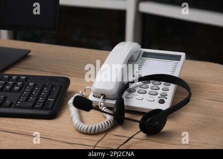 Téléphone fixe et casque près d'un ordinateur moderne sur un bureau en bois à l'intérieur. Service d'assistance téléphonique Banque D'Images