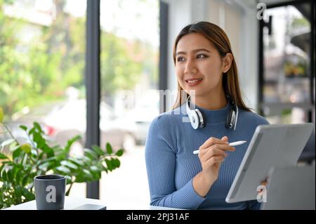 Bonne femme asiatique millénaire assis dans le café avec sa tablette portable regardant par la fenêtre, sondant et rêvant de sa vie heureuse. Banque D'Images