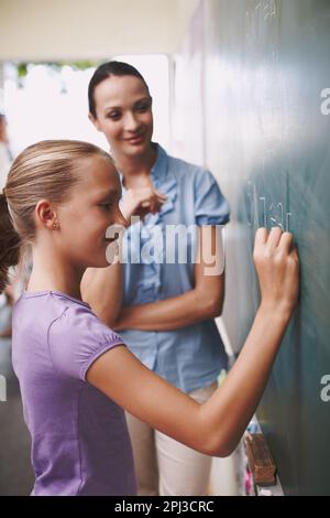 Les mathématiques sont importantes pour son avenir. Une jeune fille faisant ses sommes sur le tableau noir pendant que son professeur supervise. Banque D'Images