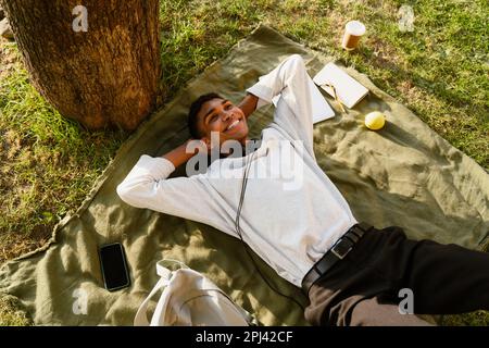 Jeune homme noir portant le sourire piercing tout en étant allongé sur une couverture sous l'arbre dans le parc Banque D'Images