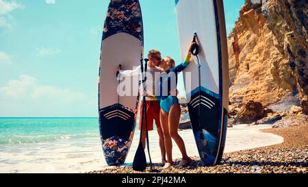Homme et femme vêtus de maillots de bain baiser sur une plage isolée tenant des planches de dessus. Jeune couple actif se tient sur une plage embrassant et tenant debout pa Banque D'Images