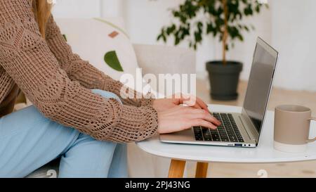 Gros plan sur un ordinateur portable de saisie des mains féminin dans le salon. Une femme d'affaires méconnue avec un ordinateur au bureau à la maison s'assoit sur un canapé près d'une table basse Banque D'Images