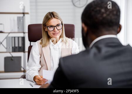 Embauche, entrevue. Femme d'affaires blonde d'âge moyen menant une interview dans un bureau lumineux, un homme afro-américain. Il regarde son CV et Banque D'Images