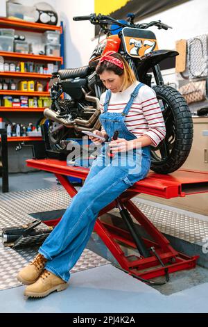 Mécanicien femme regardant le téléphone et tenant la bière assis sur la plate-forme avec moto à l'usine Banque D'Images