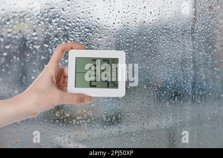 Femme tenant un hygromètre numérique avec thermomètre près de la fenêtre le jour de la pluie, gros plan. Espace pour le texte Banque D'Images