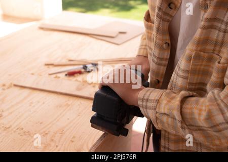 Vue latérale de la récolte un homme non reconnaissable coupe une planche en bois avec scie sauteuse électrique sur établi tout en fixant des meubles dans la salle de travail Banque D'Images