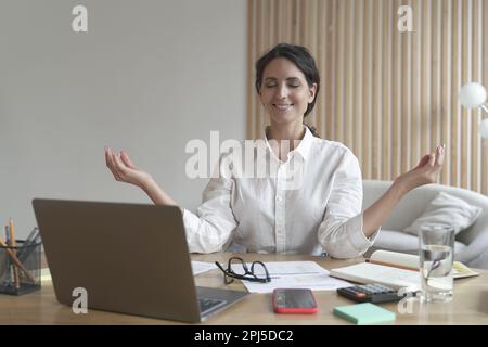 Calme paisible femme freelance méditant, pratiquant le yoga au travail tout en étant assis dans la posture zen au bureau à domicile, souriant femme d'affaires heureuse faisant des respirent Banque D'Images