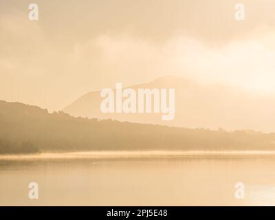 Une paisible scène matinale d'un lac brumeux reflétant les montagnes ensoleillées et les arbres, entouré par la nature intacte. Banque D'Images