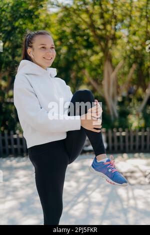 La coupe verticale de la femme énergique mène un style de vie actif garde le genou levé exercices en plein air vêtu d'une tenue active soulève les sourires de jambe écoute agréablement Banque D'Images