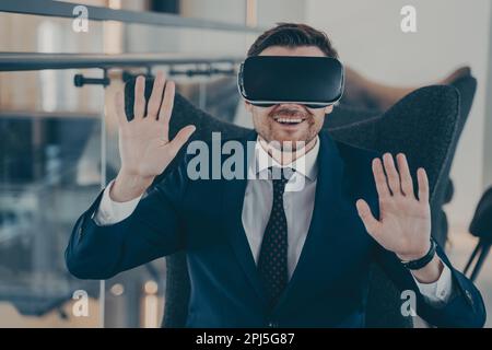 Jeune homme d'affaires heureux en costume noir formel, assis dans l'intérieur du bureau à l'aide d'un casque VR et interagit avec la simulation de réalité virtuelle, fonctionne à digit Banque D'Images