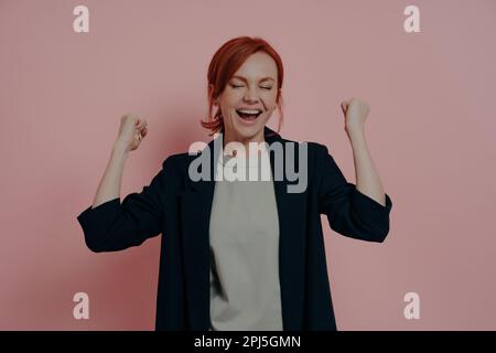 Jeune femme élégante aux cheveux rouges vêtue d'une veste marine décontractée hurlant de l'excitation avec les yeux fermés et les poings serrés, atteignant le but, celebratio Banque D'Images