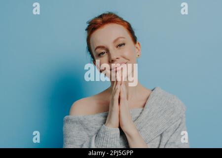 Calme belle jeune femme avec les cheveux rouges dans un petit pain faisant le souhait avec les paumes pressés, priant dieu, demandant la bonne chance tout en se tenant contre le mur bleu, re Banque D'Images