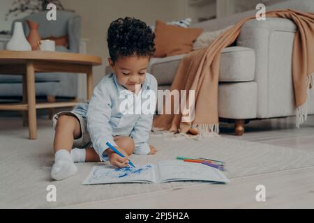 Adorable enfant africain assis sur le sol à la maison dans un salon élégant avec des stylos feutres de couleur et dessin dans un album avec l'expression concentrée sur sa fa Banque D'Images