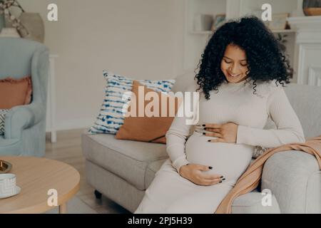 Bonne jeune femme afro-américaine en robe blanche avec de longs cheveux bouclés regardant son ventre avec un sourire doux, prenant peu de pause des travaux ménagers Banque D'Images