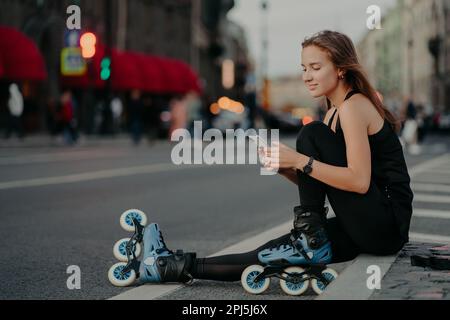Photo en extérieur d'une jeune femme sportive active vêtue de vêtements actifs porter des rollerblades vérifie le contenu des messages sur les poses de smartphone contre la ville floue ba Banque D'Images