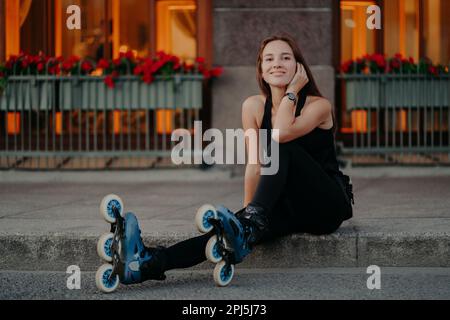 La photo horizontale d'une jeune femme active prend une pause tandis que les roussettes dans la rue vêtues de vêtements de sport noirs se sent sourires satisfaits pose agréablement Banque D'Images