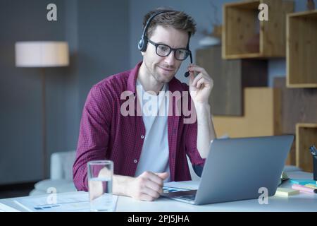 Beau sourire autrichien homme dans le casque avec micro parlant par appel vidéo à la maison, sympathique homme agent de centre d'appel consultant client en ligne usin Banque D'Images