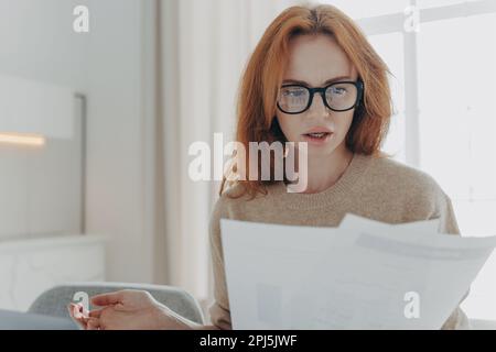 Sérieuse redhead jeune femme concentrée dans des documents papier études rapport financier porte des lunettes décontracté pull pose à l'intérieur contre l'intérieur de la maison. Bus Banque D'Images