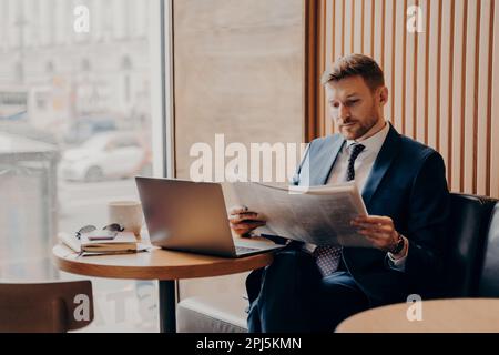 Travailleur d'entreprise masculin concentré en costume bleu formel assis dans le coin du café à côté de la grande fenêtre, lisant le journal de près, travail ouvert lapto Banque D'Images