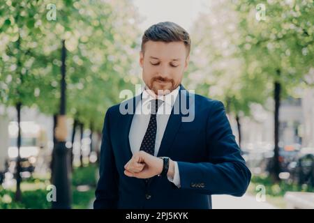 Jeune homme d'affaires attrayant avec une barbe bien taillée dans un costume moderne et élégant, attendant la date de la réunion dans le parc en plein air, l'été ensoleillé, avec impatience Banque D'Images