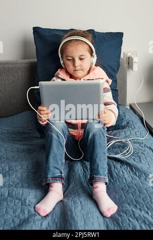 Petite fille preschooler apprendre en ligne résoudre des puzzles jouant des jeux éducatifs listetning à la musique et des sons en utilisant la tablette et les headpphones à la maison Banque D'Images