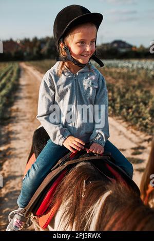 Petite fille souriante apprenant l'équitation. 5-6 ans équestres en casque ayant plaisir à monter un cheval Banque D'Images