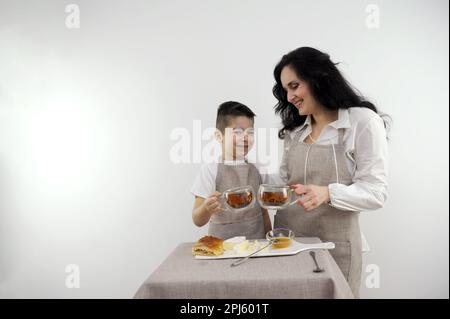 maman et petit fils s'étaler sur le fromage miel petit déjeuner nourriture saine sur fond blanc boire thé tablier de cuisine Banque D'Images