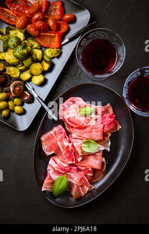 Prosciutto avec différents hors-d'œuvre avec un verre de vin rouge sur fond noir. Sur les légumes rôtis, en-cas sain Banque D'Images