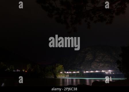 Le pont piétonnier traversant la rivière Katun brille de lanternes sous une montagne la nuit en Altai, en Sibérie, derrière des branches d'arbres. Banque D'Images