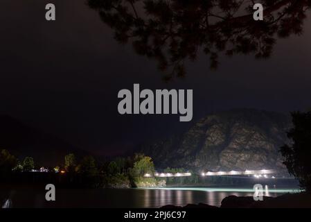 Le pont piétonnier traversant la rivière Katun brille de lanternes sous la montagne la nuit dans l'obscurité de l'Altaï en Sibérie, derrière les branches d'un arbre Banque D'Images