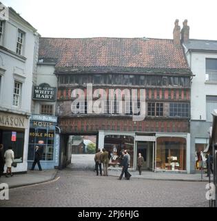 Vers la fin de 1960s, vue historique, exterrifiante de cette époque, debout sur une rue pavée, deux hommes regardant un vieux bâtiment, Ye Olde White Hart au sud-est de la place du marché, Newwark on Trent, Angleterre, Royaume-Uni. Cette ancienne auberge de coaching avec entrée voûtée date du 14th siècle. Sur la gauche, à côté de Ye Olde Pie Shoppe, vendant des Noble Pies. Au-dessous de la vieille auberge, un shope, Bainbridges. Boutique à gauche de Ye Olde White Hart, Mason, avec une affiche promotionnelle pour le beurre de kangourou, 3/- ( 3 shillings) dans la fenêtre, Banque D'Images