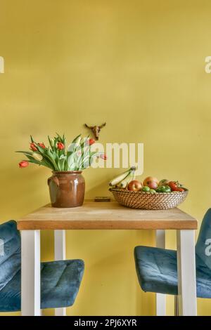 deux chaises et une table avec des fleurs au milieu, sur un mur jaune derrière elle est un panier de fruits Banque D'Images