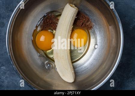 Bol en métal avec œufs, banane mûre et poudre de cacao pour faire des crêpes à la banane. Nourriture drôle en forme de visage, gros plan Banque D'Images