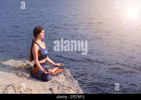 Jeune fille méditant sur la falaise près de la rivière au lever du soleil. Pratiquer le yoga Banque D'Images