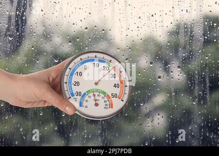 Femme tenant un hygromètre avec thermomètre près de la fenêtre le jour de la pluie, gros plan Banque D'Images