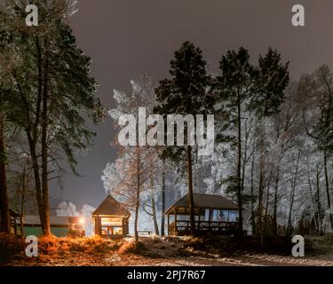 Une maison de vacances sur la rive avec deux femmes qui se reposent dans la neige sous des birches la nuit avec un feu de brasier à Altaï dans l'obscurité. Banque D'Images