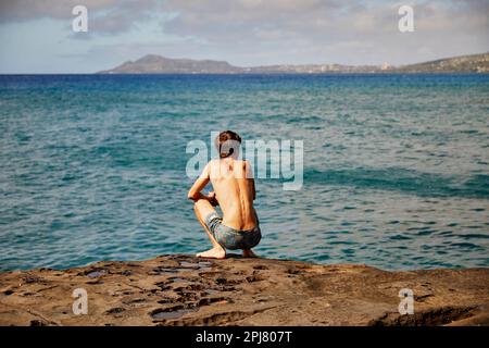 Oahu, Hawaï, États-Unis, - 7 février 2023 : un jeune surfeur qui s'accroupille sur la roche volcanique et regarde les vagues sur la côte rocheuse de l'est d'Oahu, Hawaï Banque D'Images