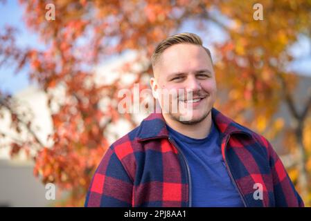 Vue de face d'un jeune homme avec un sourire éclatant dans une veste paysanne se dresse devant un fond d'automne avec des arbres et des feuilles jaunes Banque D'Images