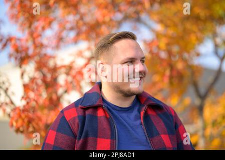 Vue latérale d'un jeune homme avec un sourire éclatant dans une veste paysanne se dresse devant un fond d'automne avec des arbres et des feuilles jaunes Banque D'Images