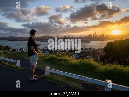 Les touristes regardent le coucher du soleil sur Auckland avec un bateau de croisière naviguant dans le port de Waitemata. Vue depuis le Mont Victoria, Devonport. Banque D'Images