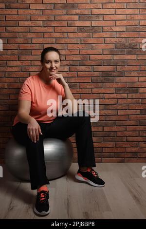Bonne femme en surpoids assise sur le ballon de fitness près du mur de briques à l'intérieur Banque D'Images