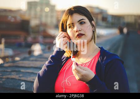 Une jeune femme de grande taille prend une pause pendant son entraînement en plein air. Elle porte un costume de jogging et on voit qu'elle porte des écouteurs filaires. Le fond sh Banque D'Images